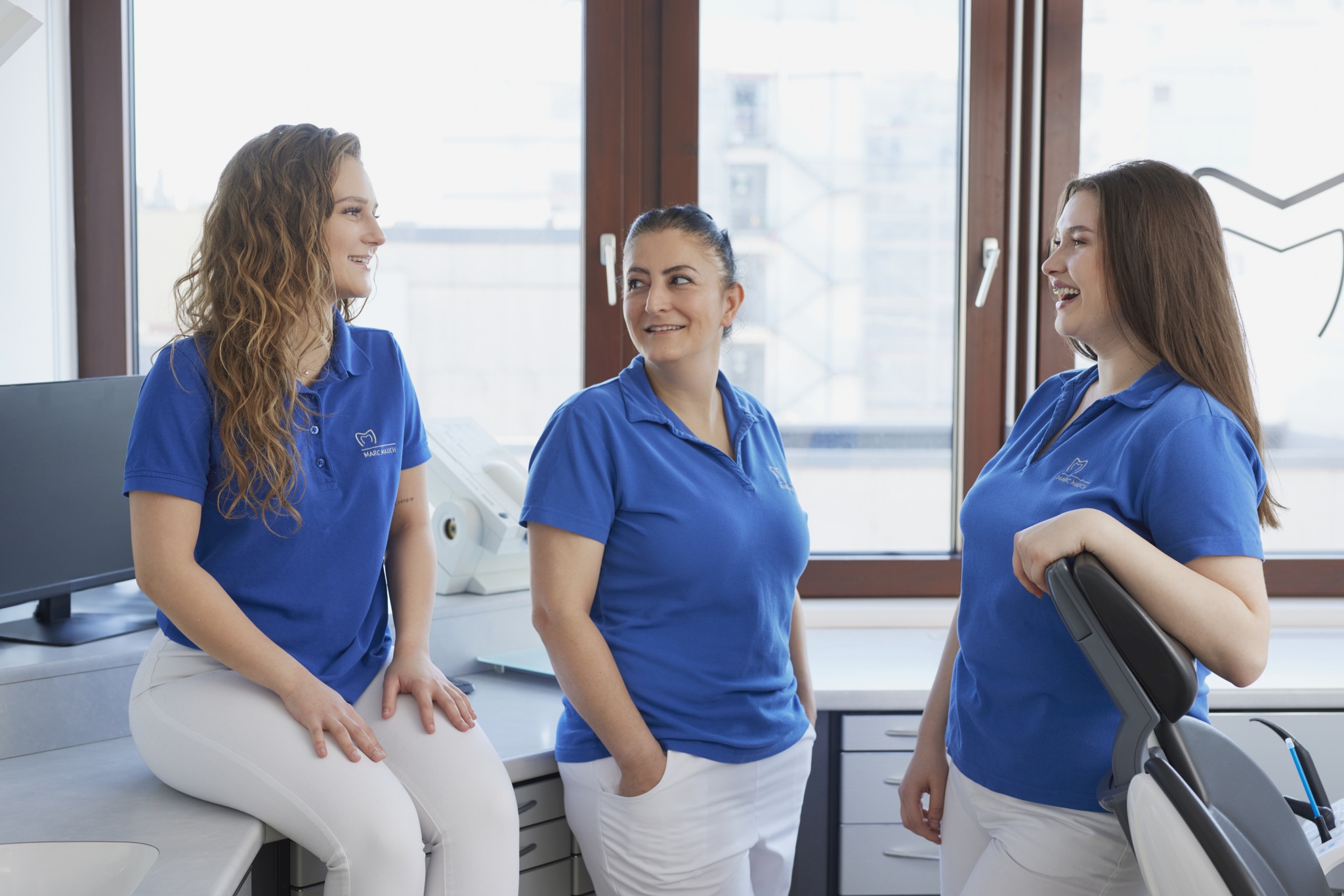 Drei Mitarbeiterinnen der Praxis in blauen Poloshirts unterhalten sich lächelnd in einem hellen Behandlungsraum; im Hintergrund Arbeitsflächen, Schränke und große Fenster.