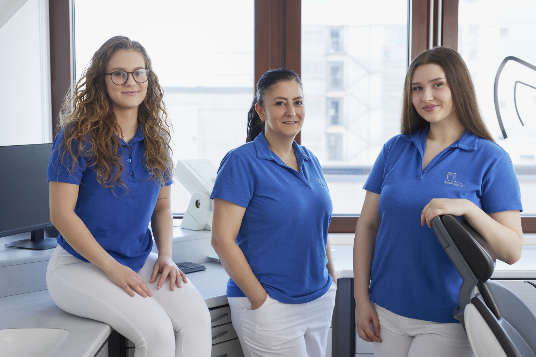 Drei Mitarbeiterinnen in blauen Poloshirts posieren nebeneinander im Behandlungsraum; hinter ihnen Arbeitsflächen, Geräte und Fensterfront mit Stadtblick.