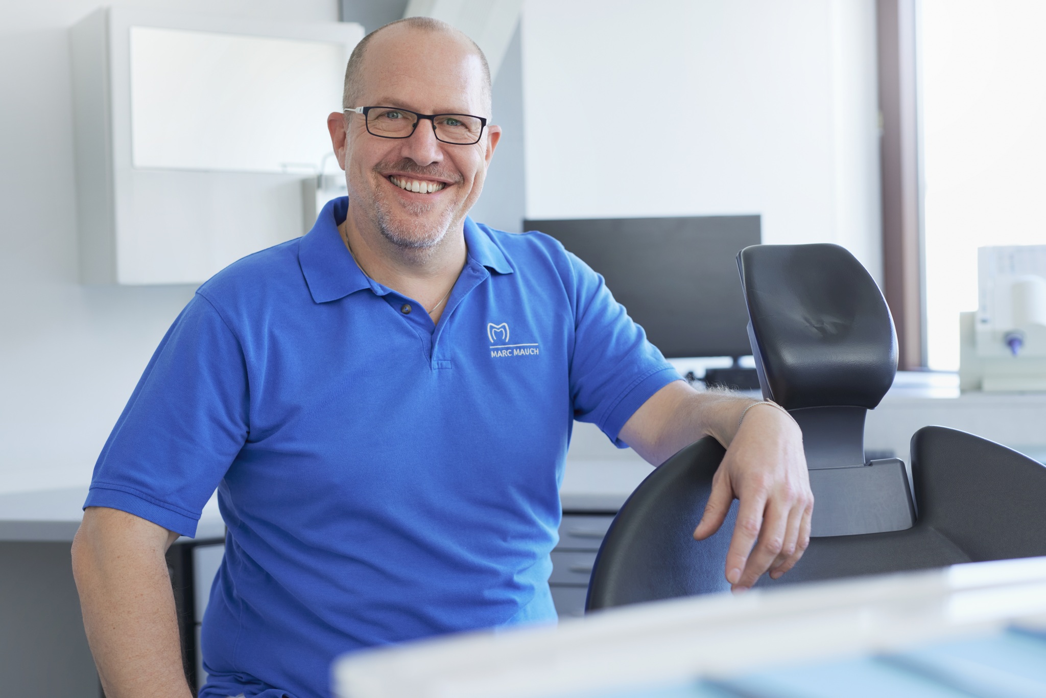 Dr. Mauch sitzt entspannt neben dem Behandlungsstuhl und lächelt; im Hintergrund Monitor, Schränke und diffuses Tageslicht.