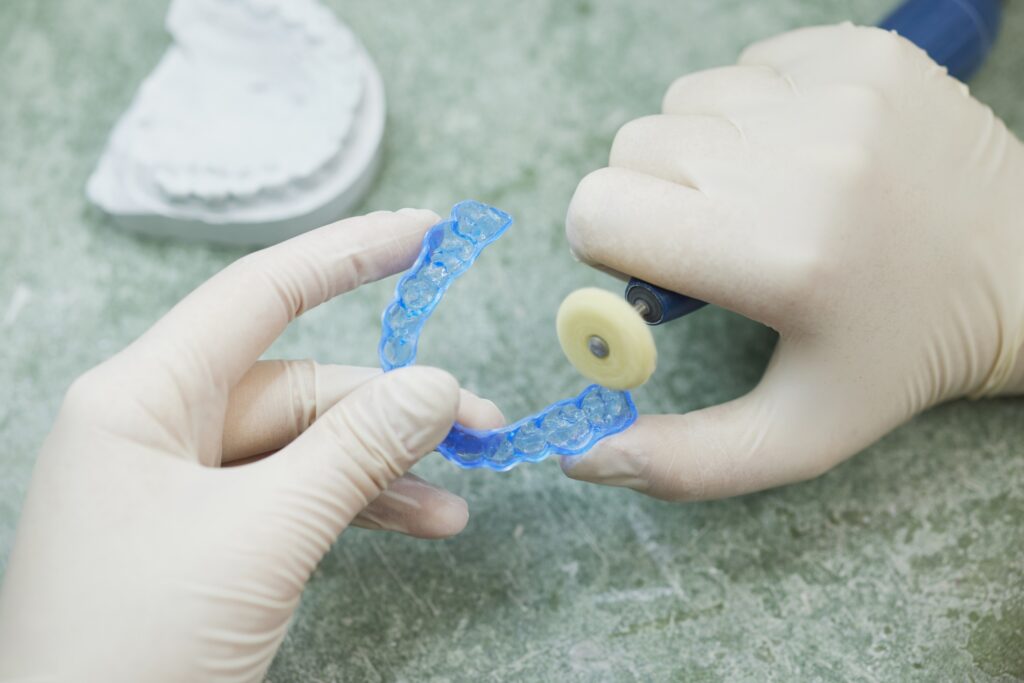 Detail: behandschuhte Hände bearbeiten eine blaue Zahnschiene mit rotierendem Instrument; grüne Arbeitsfläche, links ein Zahnabdruck.