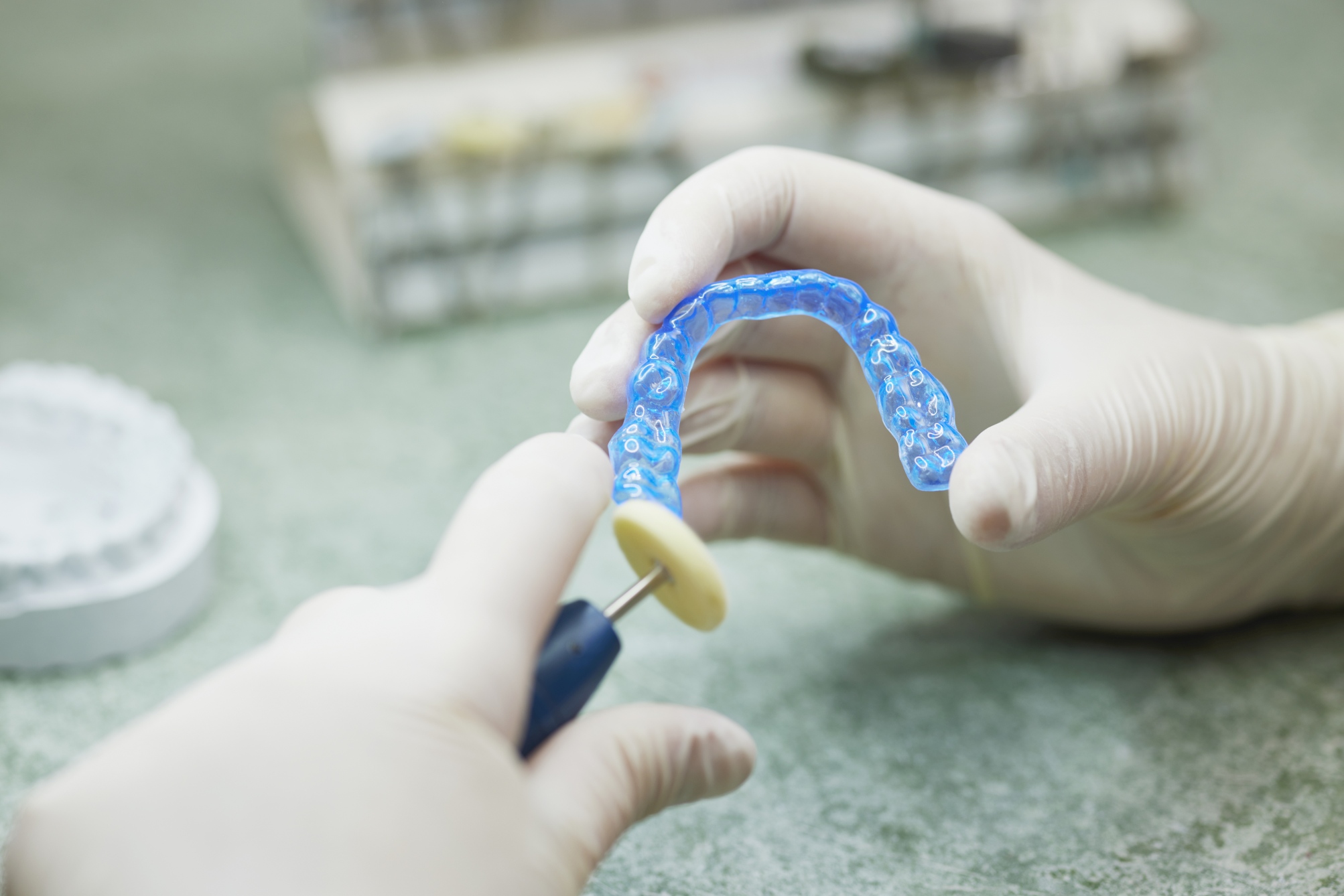 Nahaufnahme: behandschuhte Hände halten eine transparente blaue Zahnschiene und schleifen Kanten; unscharfer Laborhintergrund.