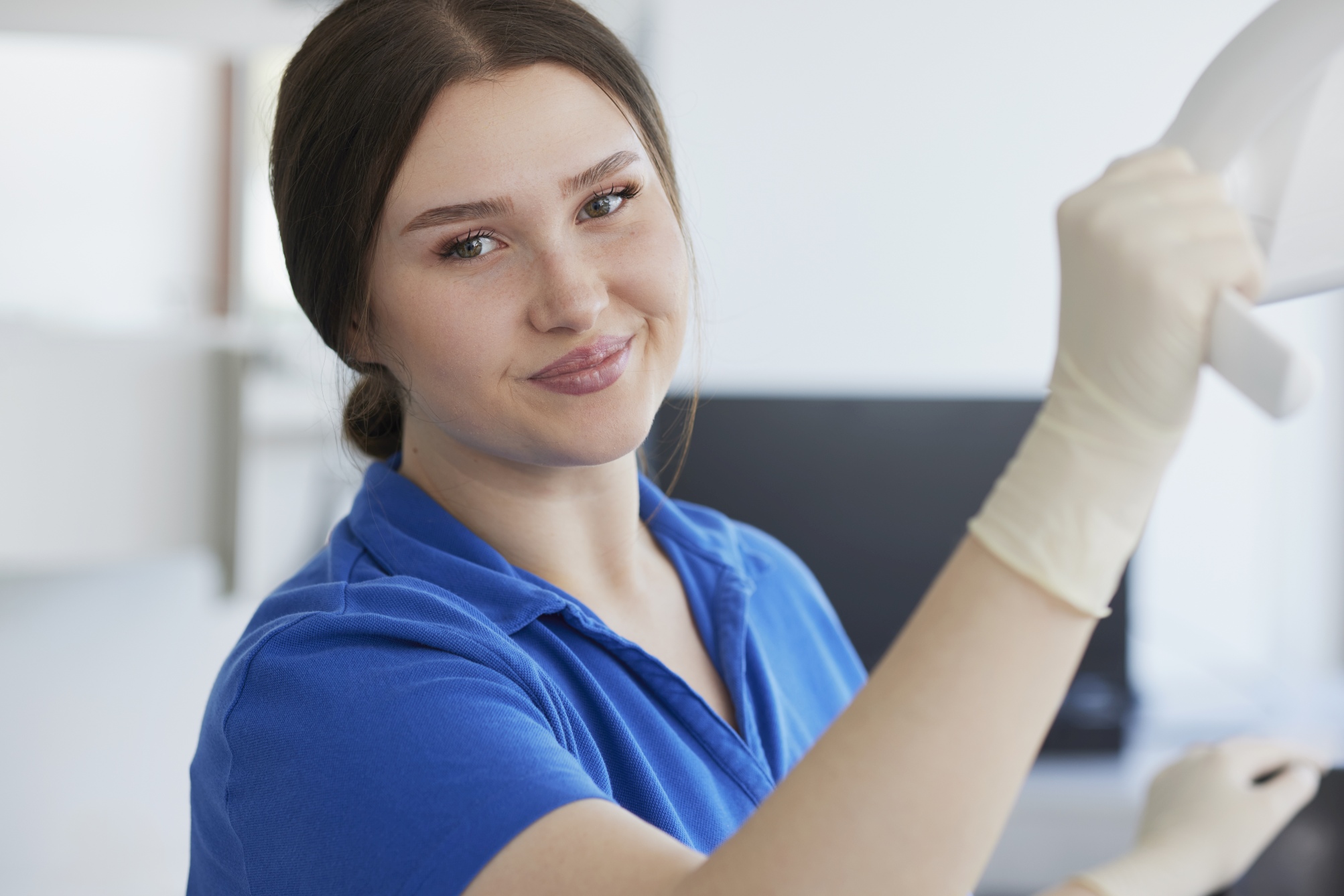 Mitarbeiterin im blauen Poloshirt hebt die Behandlungslampe an und blickt in die Kamera; dahinter Monitor und helle Praxiseinrichtung.