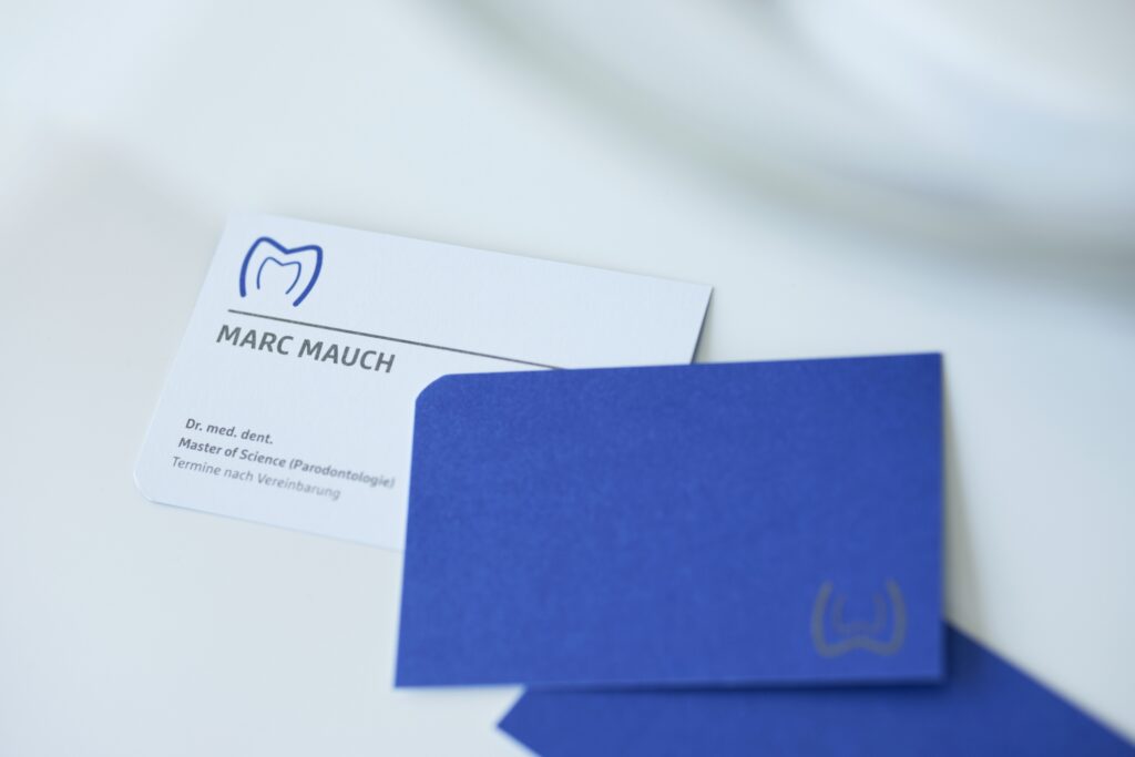 Nahaufnahme einer Visitenkarte von Dr. Marc Mauch mit Zahnlogo; daneben blauer Umschlag auf heller Oberfläche.