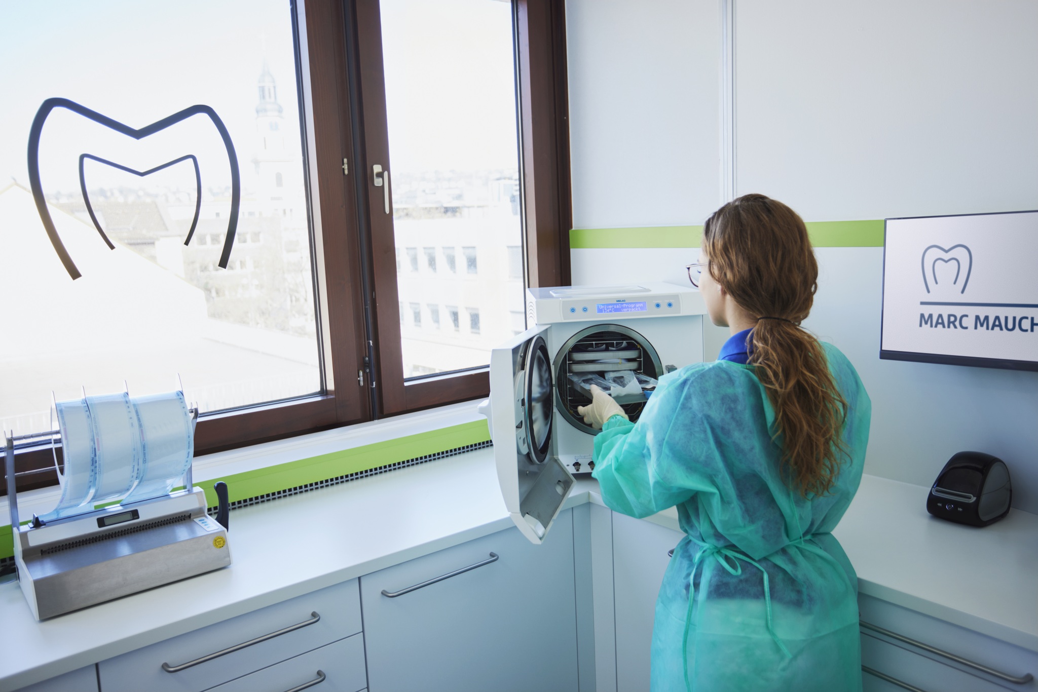 Mitarbeiterin in Schutzkittel und Handschuhen bestückt einen Sterilisator; Raum mit Fenster, Arbeitsplatte und Monitor mit Praxislogo.