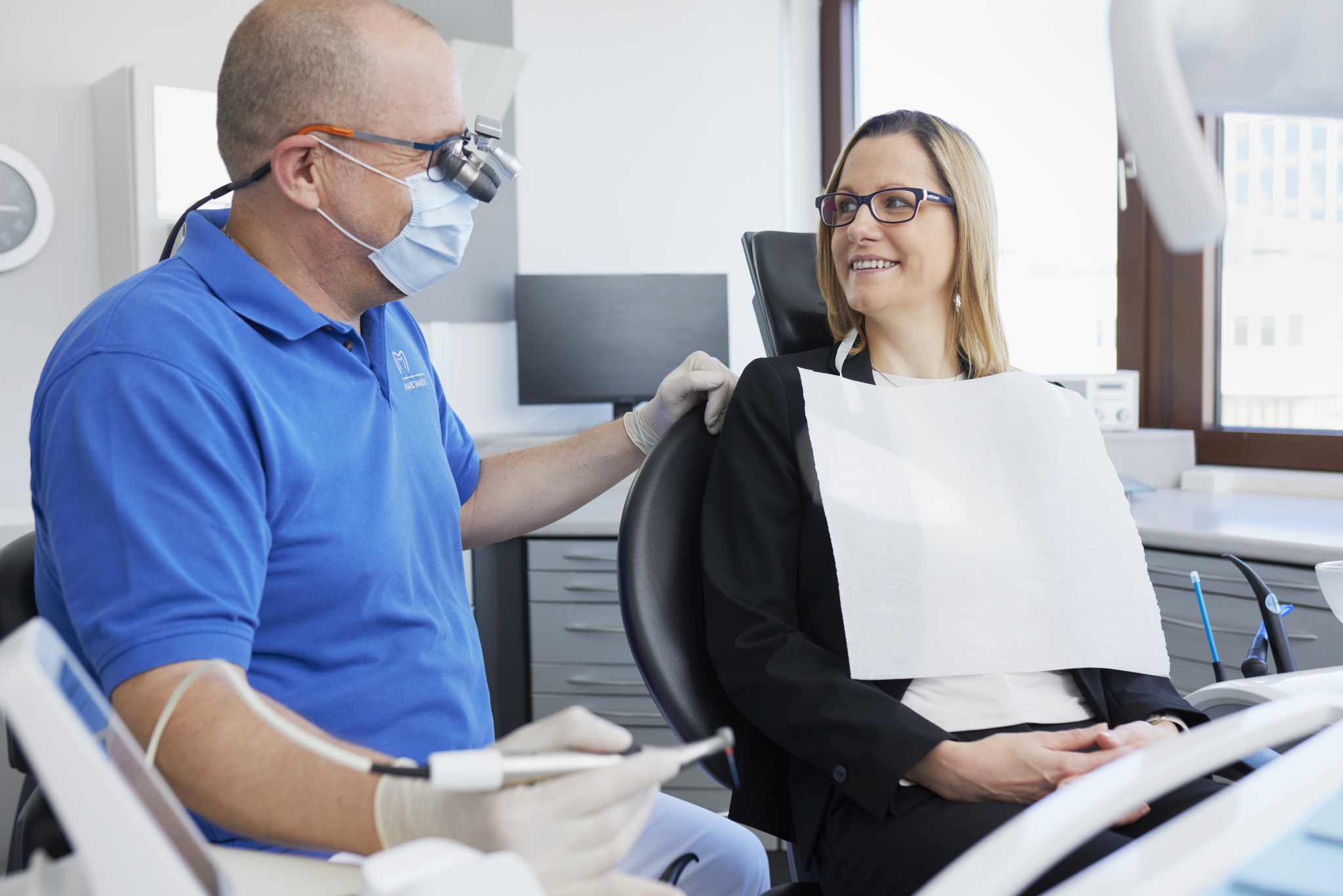 Dr. Mauch spricht mit einer Patientin im Behandlungsstuhl; helles Zimmer mit Fenstern, Instrumenten und Schränken, beide lächeln einander an.