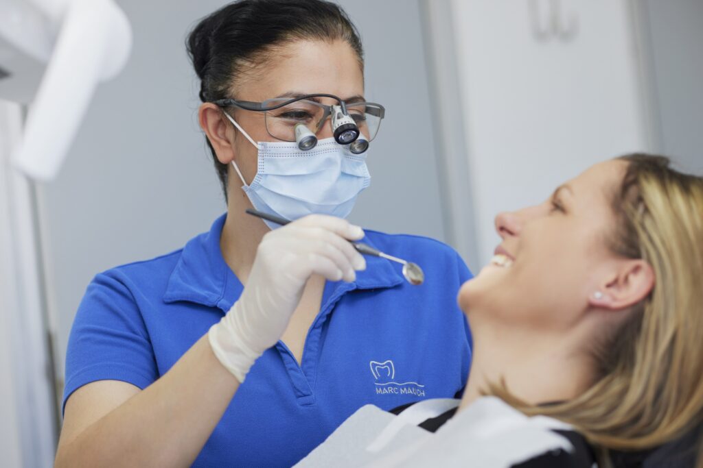 Zahnärztin mit Mund-Nasen-Schutz und Lupenbrille untersucht Patientin mit Mundspiegel; neutraler Behandlungsraum im Hintergrund.