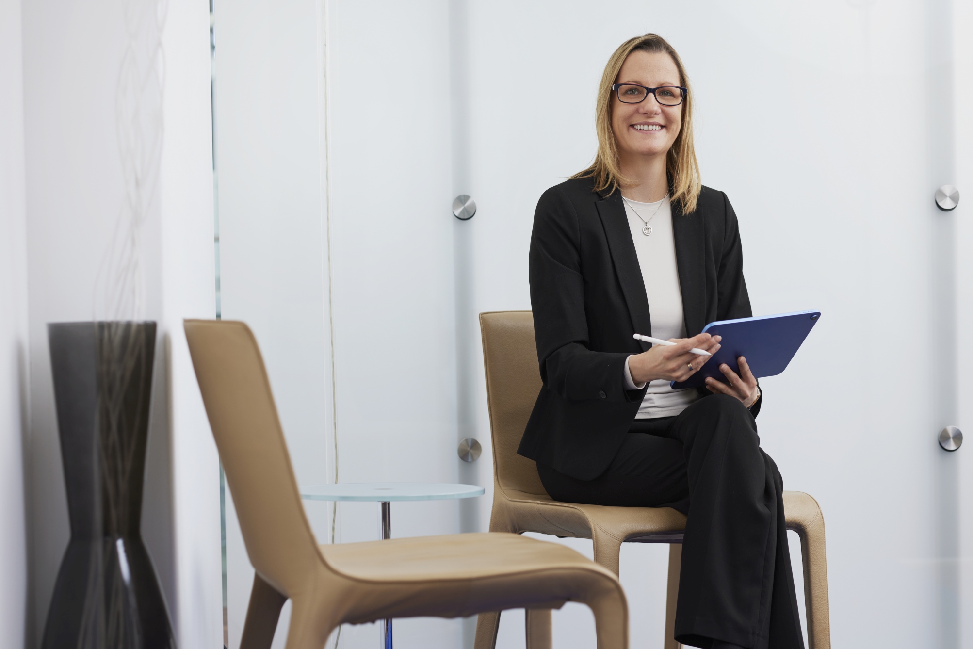 Patientin im schwarzen Blazer sitzt im Wartebereich und hält ein Tablet mit Stift; helle Glaswand und zwei Stühle im Hintergrund.
