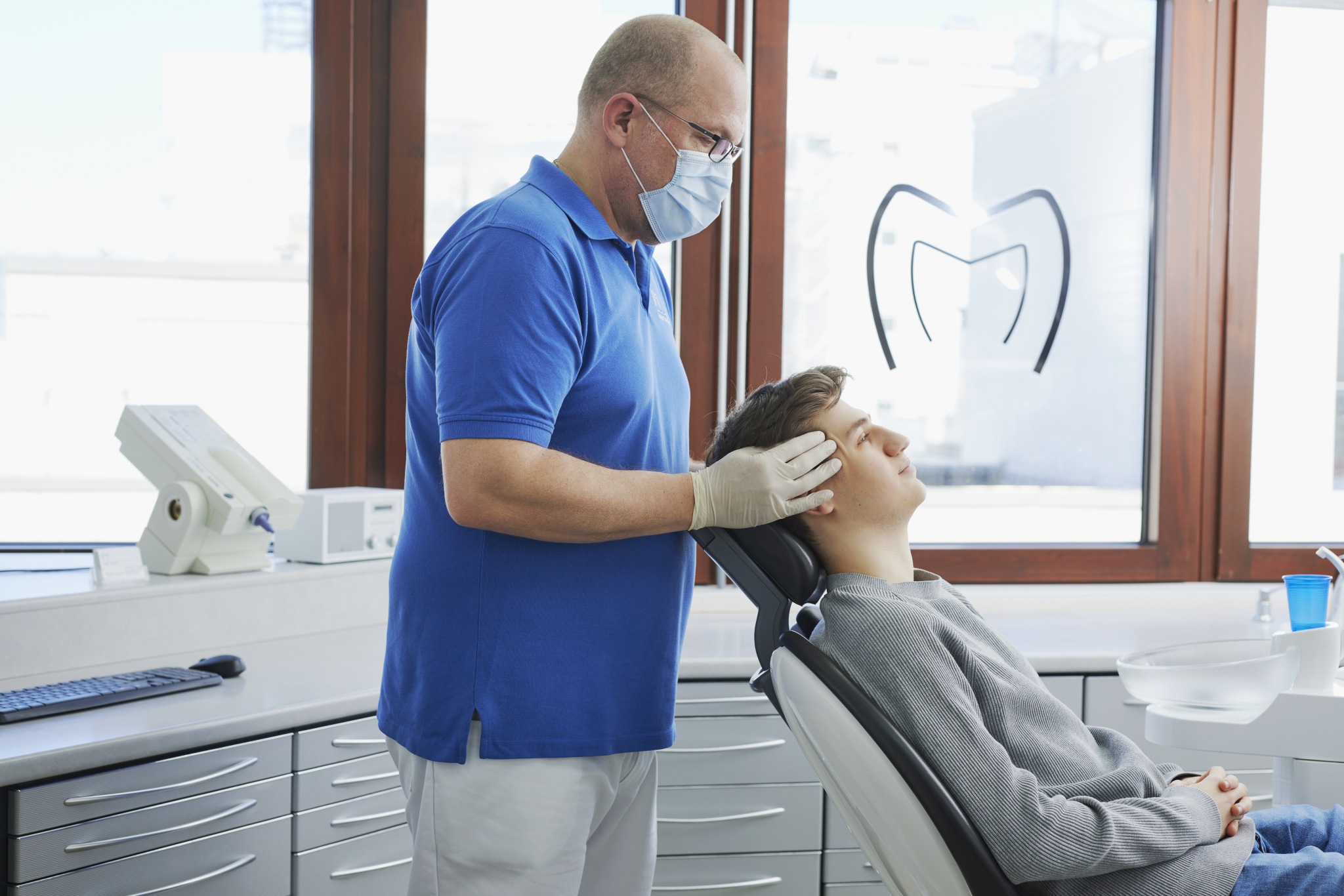 Dr. Mauch tastet den Kopf eines jungen Patienten im Behandlungsstuhl ab; große Fenster mit Praxislogo, Arbeitsflächen und Spülbecken im Hintergrund.