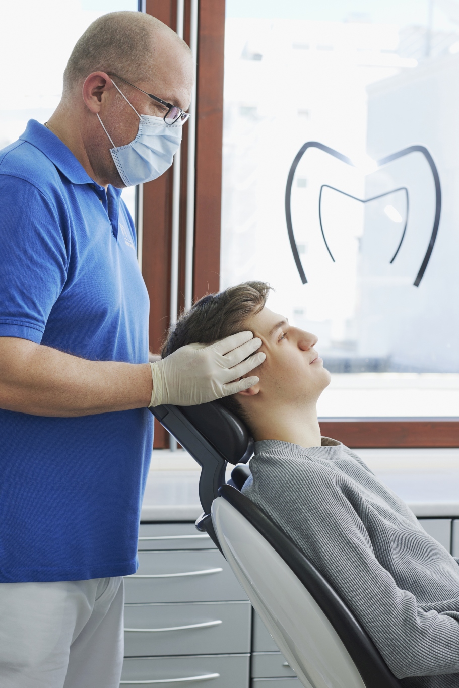 Dr. Mauch mit Mundschutz tastet die Schläfenregion eines Patienten im Behandlungsstuhl; hinter ihnen große Fenster mit Praxislogo.