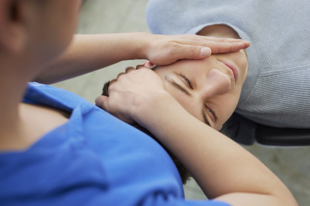 Nahaufnahme: Patient liegt im Stuhl, Hände einer Person im blauen Poloshirt stützen sanft Schläfen und Stirn; unscharfer Praxisboden im Hintergrund.