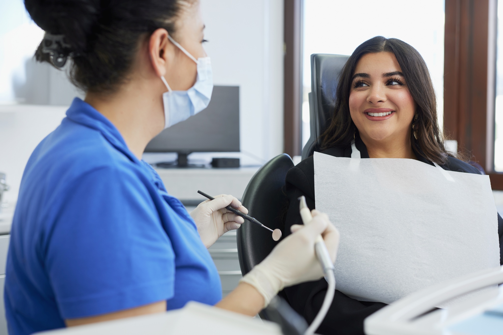 Patientin mit Schutzserviette sitzt im Behandlungsstuhl und lächelt; Zahnärztin mit Mundschutz hält Spiegel und Instrument, helles Zimmer mit Fenster.