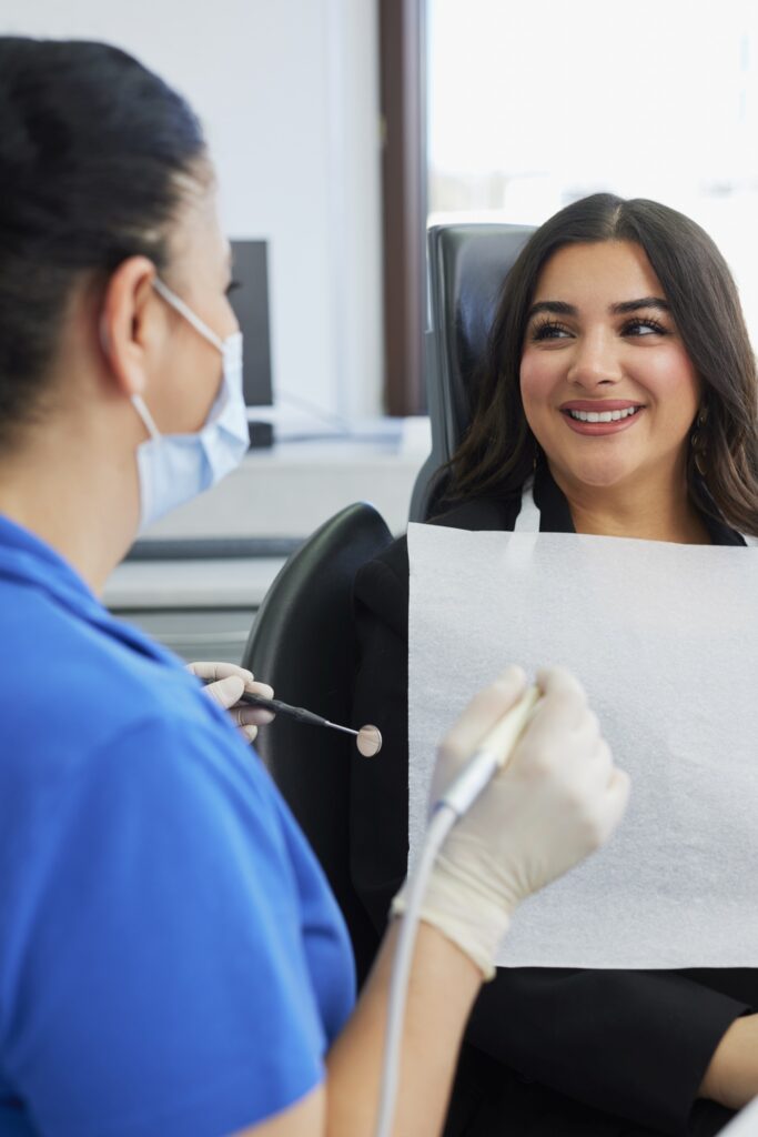 Patientin lächelt im Behandlungsstuhl, Zahnärztin mit Mundschutz hält ein Instrument; im Hintergrund Schränke und Geräte.