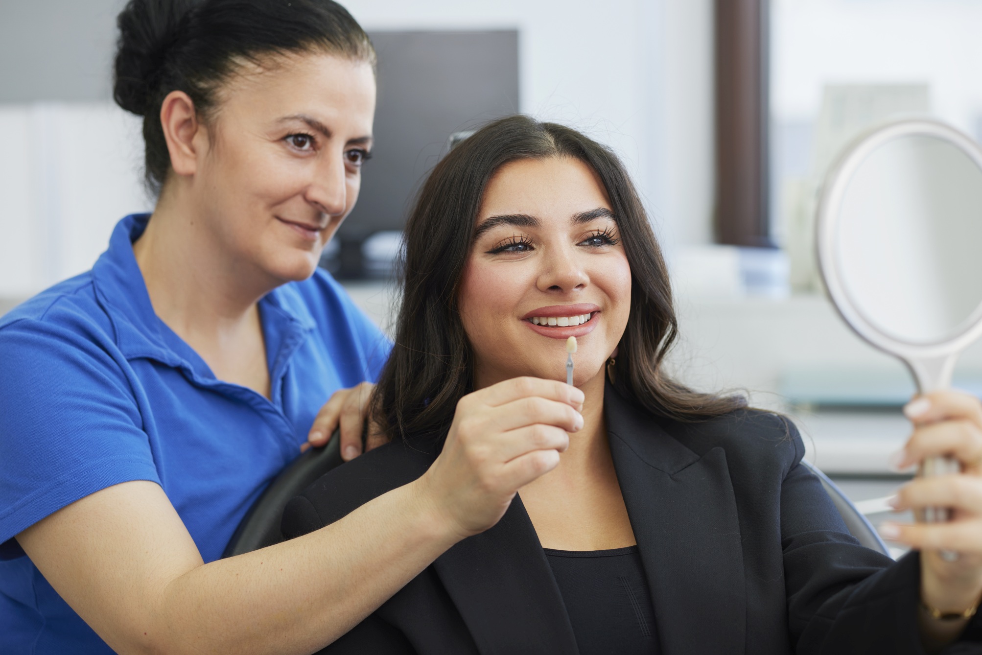 Zahnärztin zeigt einer Patientin im Stuhl mithilfe eines Handspiegels eine Zahnfarb-Probe; neutraler, heller Behandlungsraum.