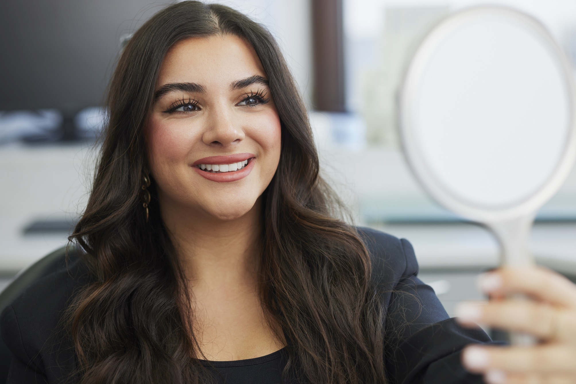 Patientin betrachtet sich lächelnd im Handspiegel; unscharfer Praxisbereich mit Monitor im Hintergrund.
