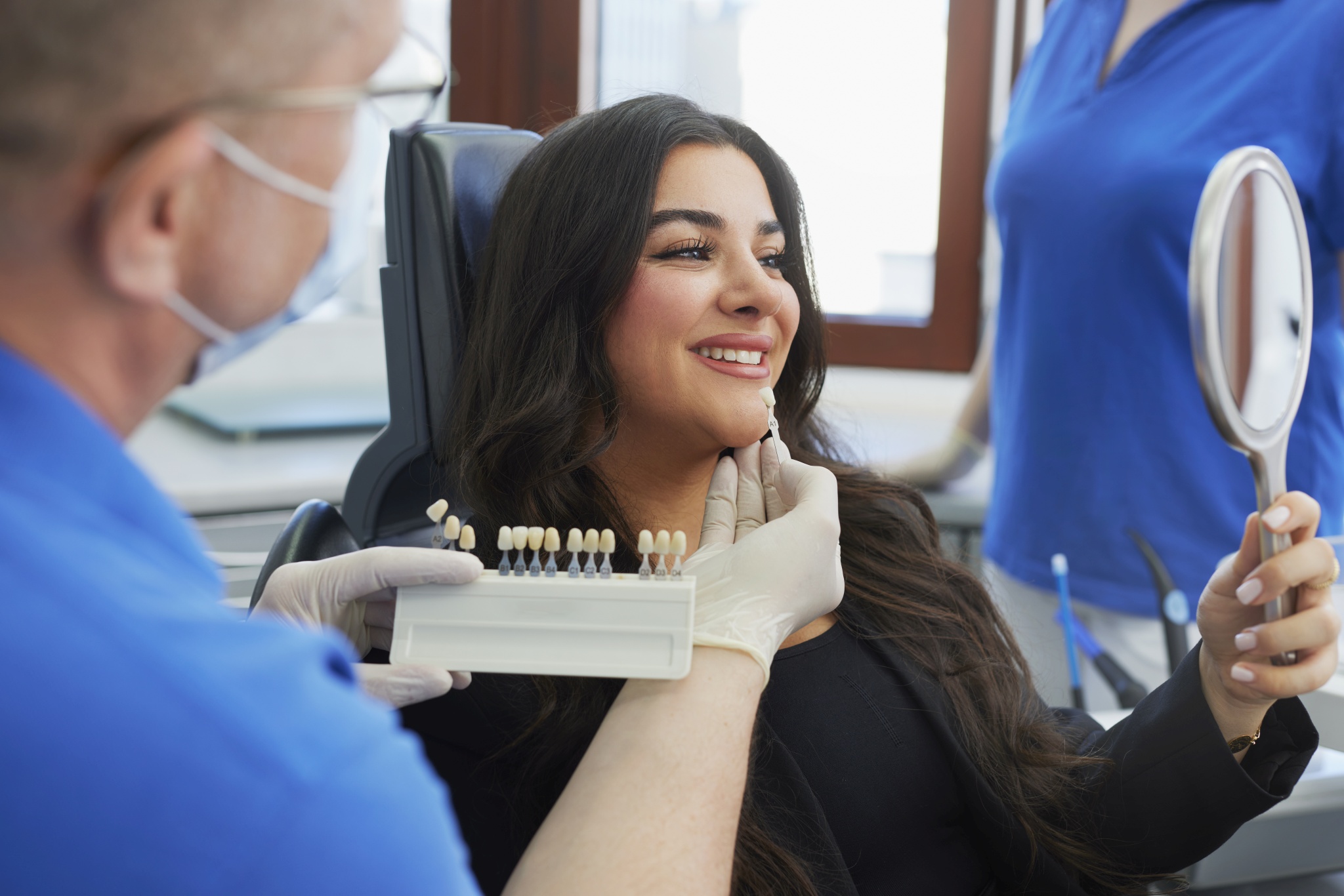 Dr. Mauch hält eine Zahnfarben-Skala an die Zähne einer Patientin, die in einen Spiegel blickt; Fenster und Behandlungsplatz im Hintergrund.
