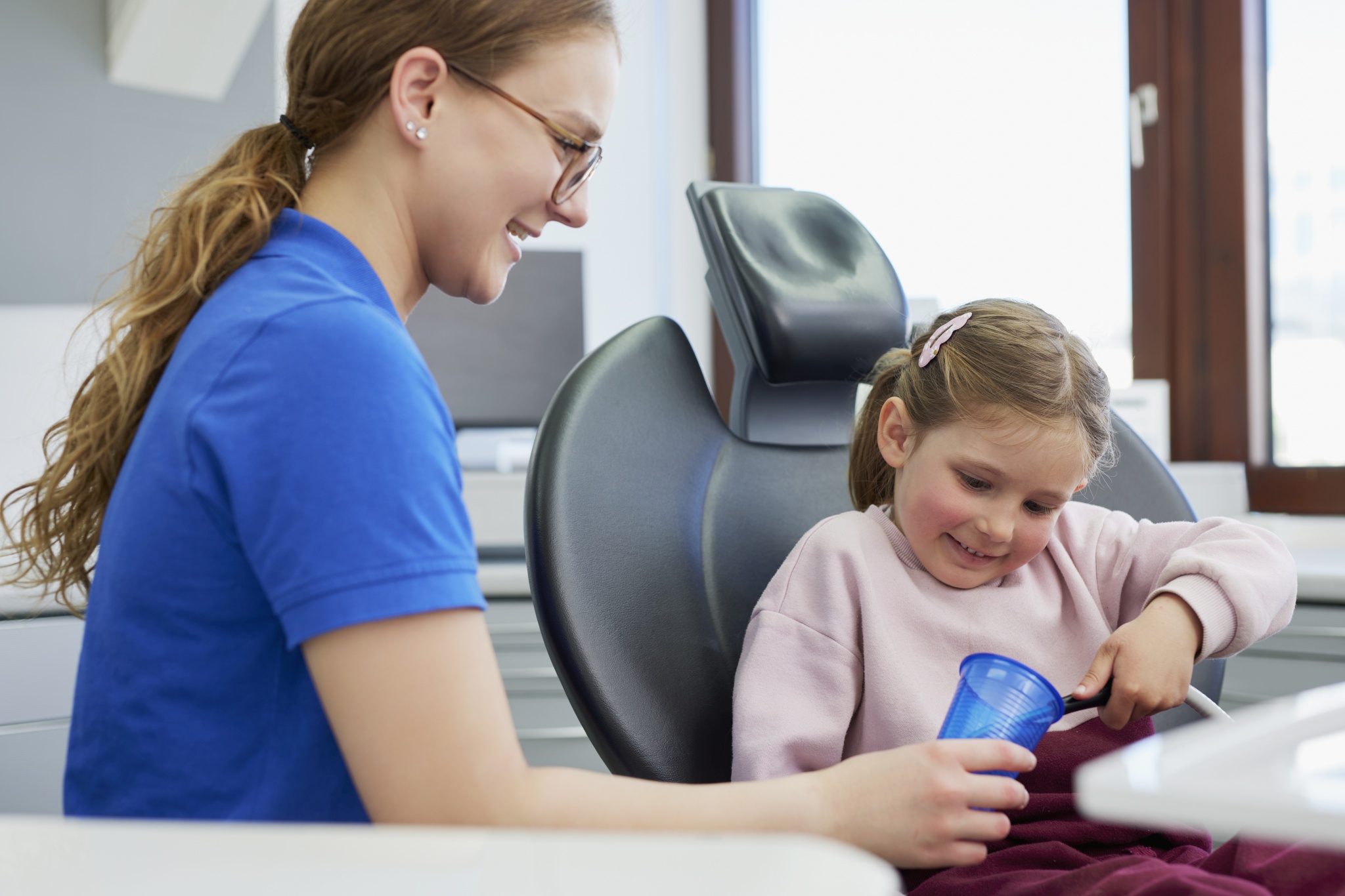 Mitarbeiterin im blauen Poloshirt sitzt neben einem Kind im Behandlungsstuhl; beide lächeln, Kind hält einen blauen Becher, Fenster im Hintergrund.