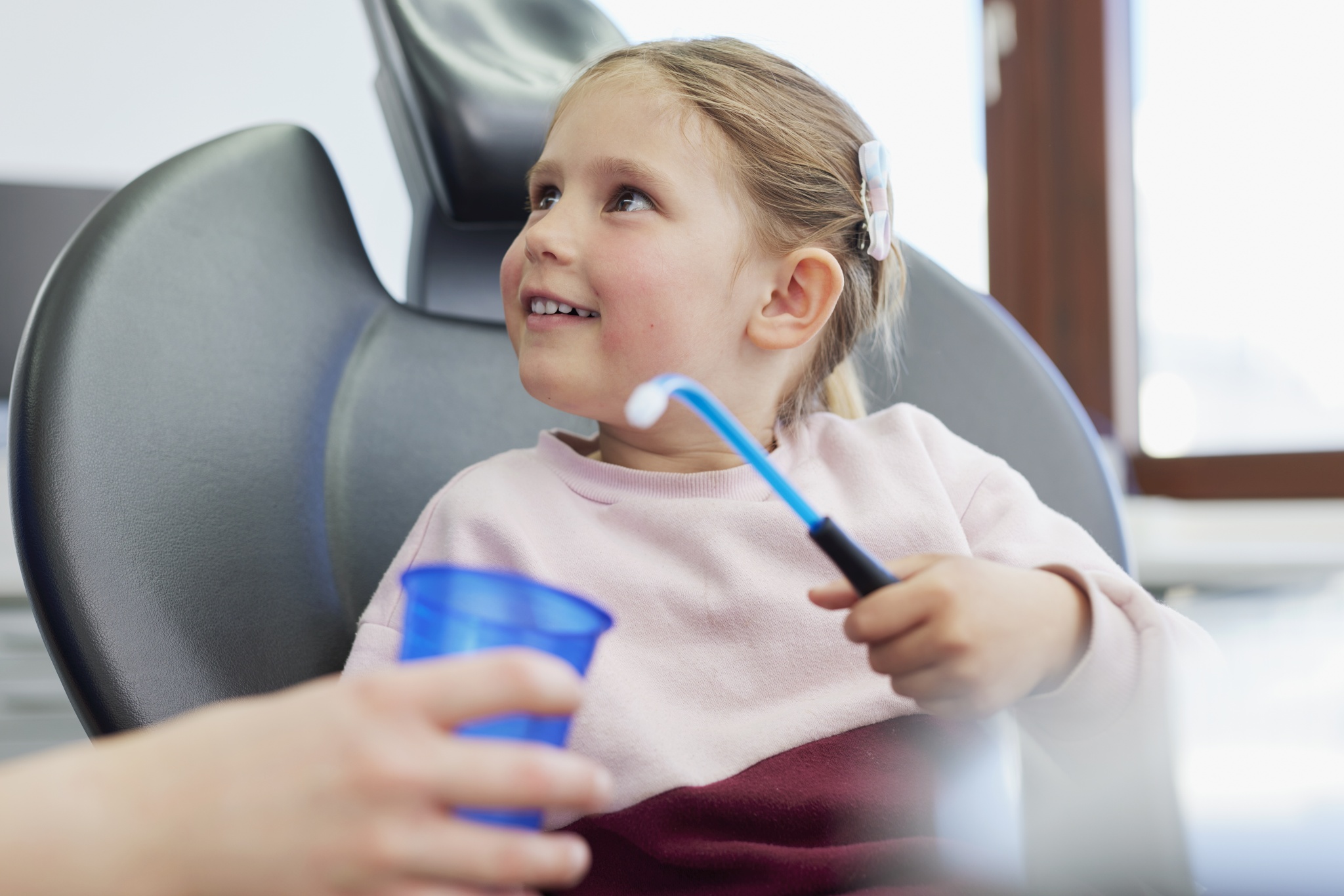 Kind im Behandlungsstuhl hält einen blauen Becher und eine Absaugkanüle; helle Praxisumgebung mit Fenster im Hintergrund.