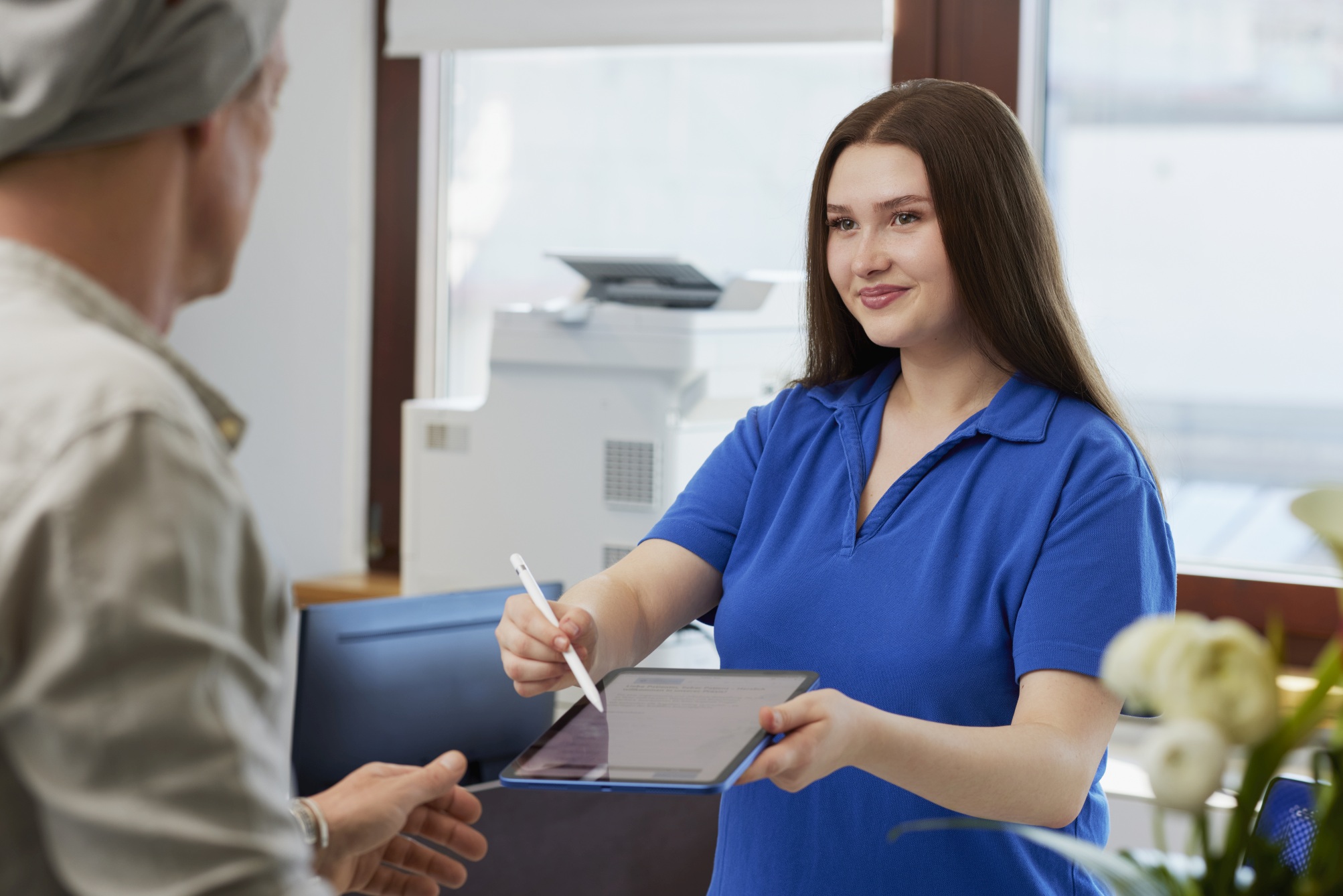 Mitarbeiterin im blauen Poloshirt reicht einem Patienten ein Tablet mit Stift zum Unterschreiben; Empfangsbereich mit Fenster, Drucker und Blumen.